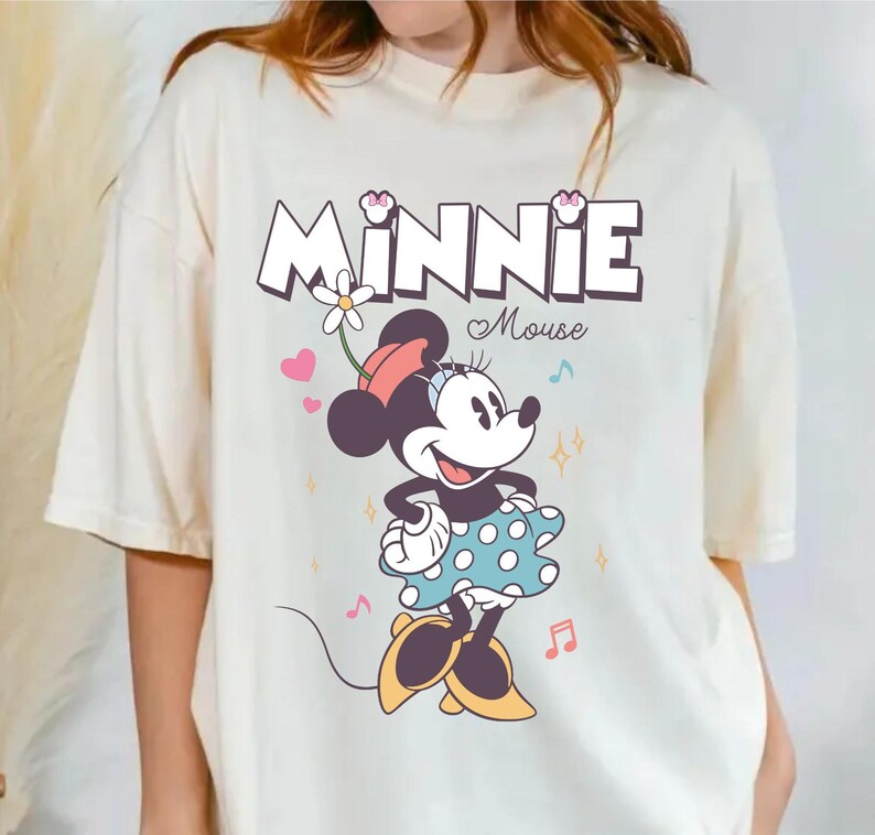 Puede incluir: Una camiseta blanca con un gr&aacute;fico de Minnie Mouse con un vestido azul de lunares y un lazo rojo. El texto "Minnie Mouse" est&aacute; impreso encima de ella en una fuente colorida.