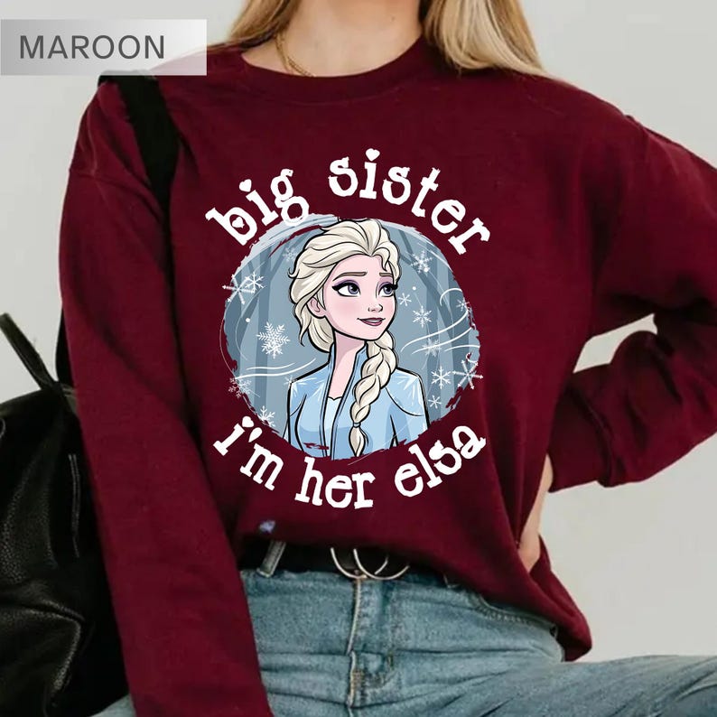 May include: Maroon sweatshirt with the text "big sister, I'm her Elsa" in white. The graphic features a cartoon image of Elsa from Frozen. The sweatshirt is made of a soft material and is suitable for casual wear.