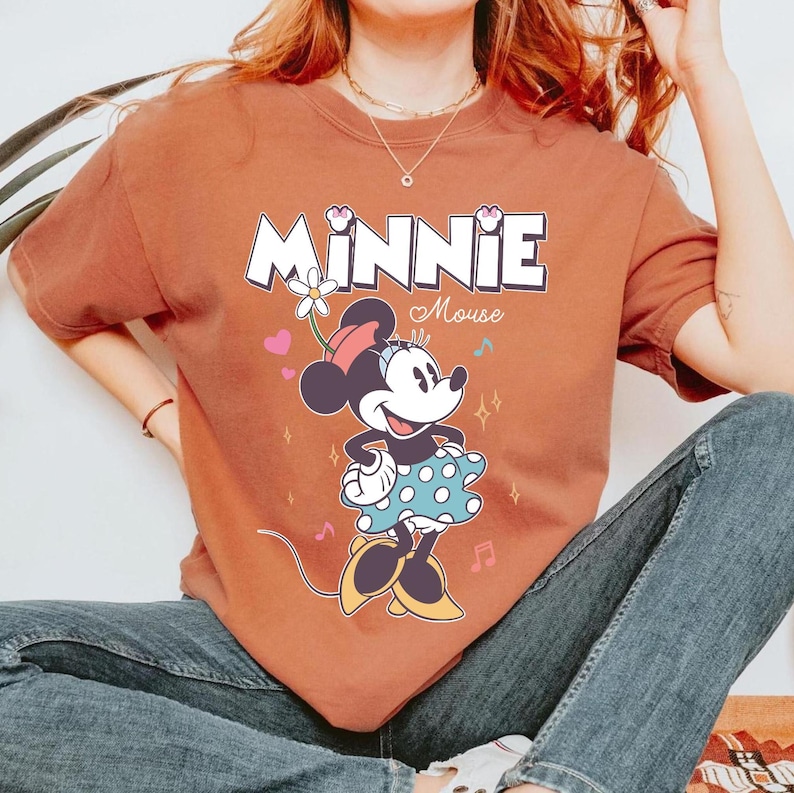 Puede incluir: Una camiseta de color &oacute;xido con un gr&aacute;fico de Minnie Mouse con un vestido de lunares azules y blancos. El texto "MINNIE Mouse" est&aacute; impreso encima de ella.