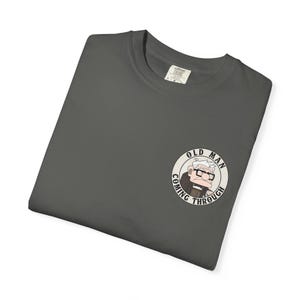 May include: Dark gray t-shirt with a circular graphic of an elderly man wearing glasses and the text "OLD MAN COMING THROUGH". The shirt is folded and has a tag on the collar.
