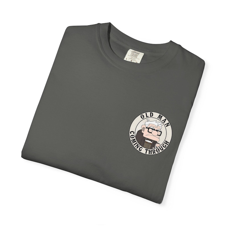 May include: Dark gray t-shirt with a circular graphic of an elderly man wearing glasses and the text "OLD MAN COMING THROUGH". The shirt is folded and has a tag on the collar.