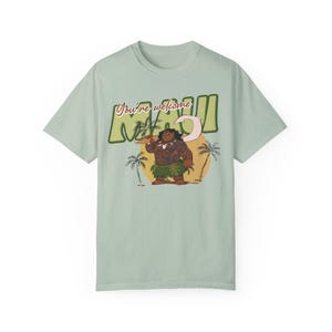 Puede incluir: Una camiseta verde claro con un estampado gr&aacute;fico de Maui de la pel&iacute;cula de Disney Moana. El gr&aacute;fico muestra a Maui sosteniendo un anzuelo y de pie frente a una puesta de sol con palmeras. El texto "You're welcome Maui" est&aacute; impreso en una fuente retro.