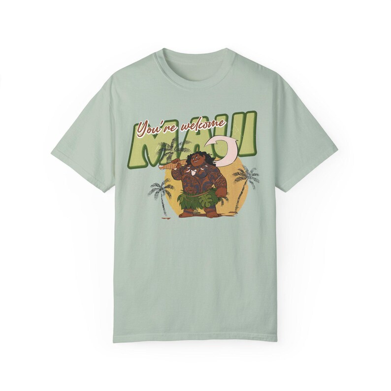 Puede incluir: Una camiseta verde claro con un estampado gr&aacute;fico de Maui de la pel&iacute;cula de Disney Moana. El gr&aacute;fico muestra a Maui sosteniendo un anzuelo y de pie frente a una puesta de sol con palmeras. El texto "You're welcome Maui" est&aacute; impreso en una fuente retro.
