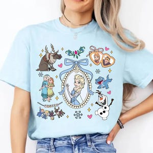 Puede incluir: Camiseta azul claro con personajes de dibujos animados de la pel&iacute;cula "Frozen". El dise&ntilde;o incluye a Elsa, Anna, Olaf, Sven y otros personajes. La camiseta tiene cuello redondo y mangas cortas.