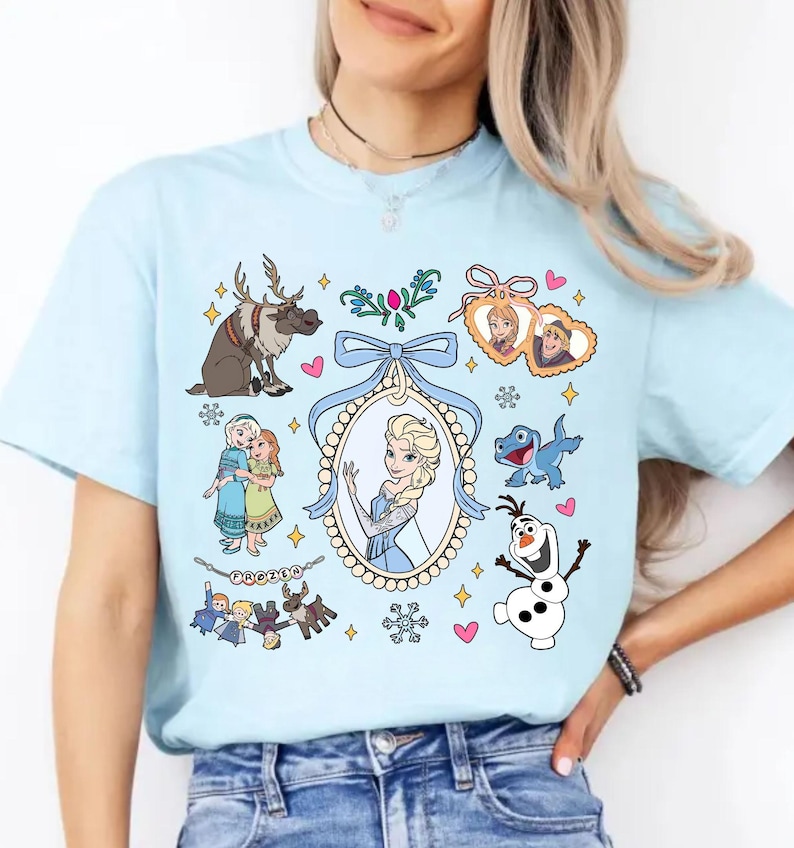 Puede incluir: Camiseta azul claro con personajes de dibujos animados de la pel&iacute;cula "Frozen". El dise&ntilde;o incluye a Elsa, Anna, Olaf, Sven y otros personajes. La camiseta tiene cuello redondo y mangas cortas.