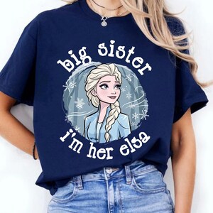 May include: Navy blue t-shirt featuring a graphic of Elsa from Frozen, surrounded by snowflakes, with the text "big sister I'm her elsa" in white. The shirt is a crew neck and is being worn.