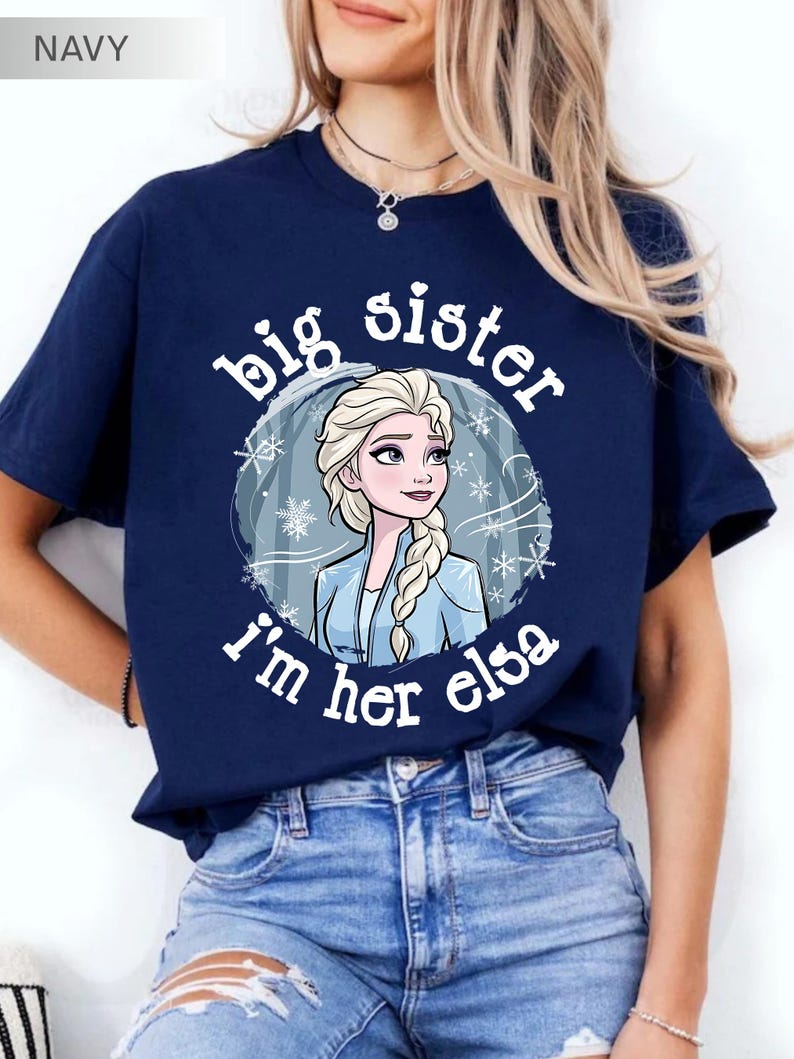 May include: Navy blue t-shirt featuring a graphic of Elsa from Frozen, surrounded by snowflakes, with the text "big sister I'm her elsa" in white. The shirt is a crew neck and is being worn.