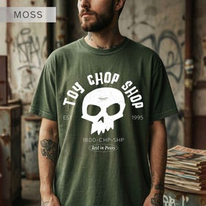 Pu&ograve; includere: Una t-shirt verde muschio con una grafica bianca raffigurante un teschio e il testo TOY CHOP SHOP, EST. 1995, 1800-CHP-SHP e Rest in Pieces. Il design presenta un aspetto usurato e invecchiato stampato sulla parte anteriore del capo.
