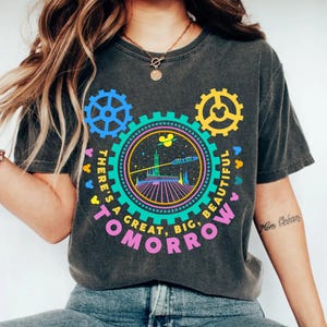 May include: A charcoal grey t-shirt featuring a colourful graphic of a futuristic space scene inside a large gear. The design includes the text "THERE'S A GREAT, BIG, BEAUTIFUL TOMORROW" in pink, with smaller gear shapes and mouse silhouettes.