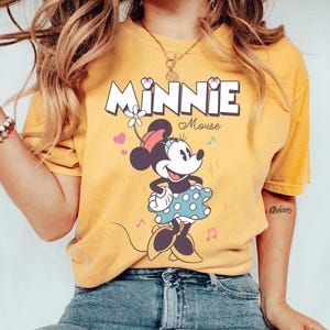 Puede incluir: Una camiseta amarilla con una imagen de dibujos animados de Minnie Mouse. La camiseta tiene el texto "Minnie Mouse" impreso.