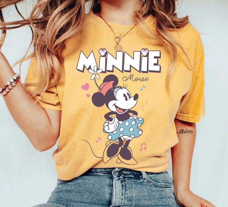 Puede incluir: Una camiseta amarilla con una imagen de dibujos animados de Minnie Mouse. La camiseta tiene el texto "Minnie Mouse" impreso.