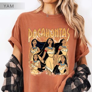 Pode incluir: T-shirt cor de ferrugem com um gr&aacute;fico de Pocahontas e a palavra "Pocahontas" em ouro. O gr&aacute;fico mostra Pocahontas com um vestido amarelo, cabelo preto comprido e um colar turquesa. A camisa tem mangas enroladas.