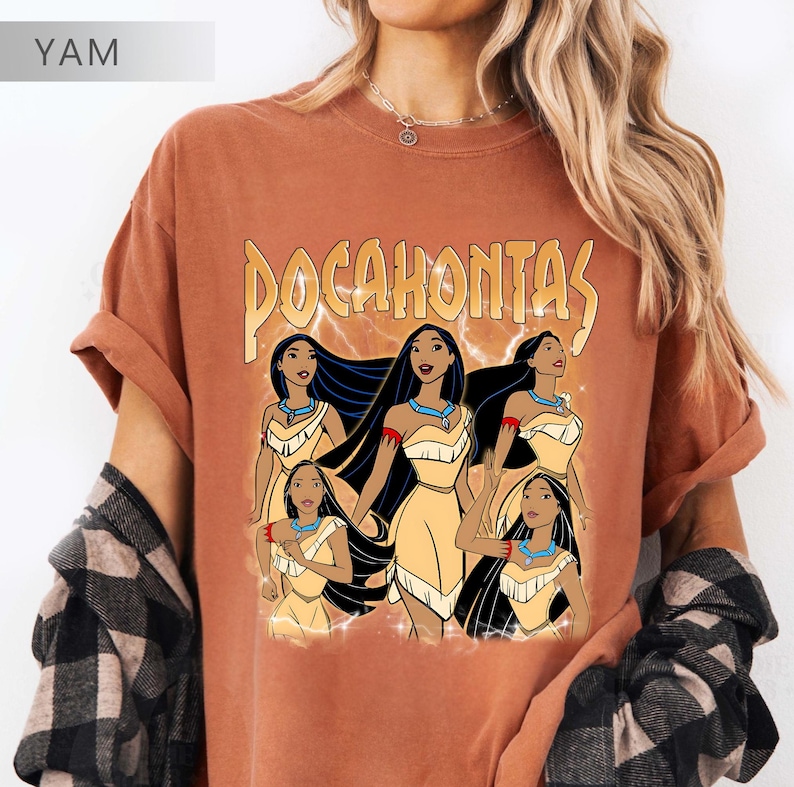 Pode incluir: T-shirt cor de ferrugem com um gr&aacute;fico de Pocahontas e a palavra "Pocahontas" em ouro. O gr&aacute;fico mostra Pocahontas com um vestido amarelo, cabelo preto comprido e um colar turquesa. A camisa tem mangas enroladas.