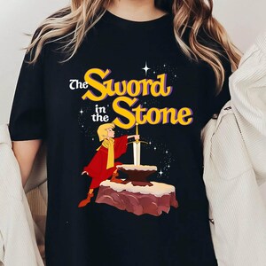 K&ouml;nnte beinhalten: Schwarzes T-Shirt mit einer Grafik aus "The Sword in the Stone". Das Design zeigt den Titel in Gelb und Lila, eine Figur in Rot und Gelb und ein Schwert in einem Stein. Das Design ist vor einem Sternenhintergrund.