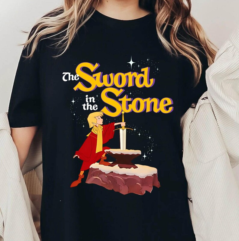 K&ouml;nnte beinhalten: Schwarzes T-Shirt mit einer Grafik aus "The Sword in the Stone". Das Design zeigt den Titel in Gelb und Lila, eine Figur in Rot und Gelb und ein Schwert in einem Stein. Das Design ist vor einem Sternenhintergrund.