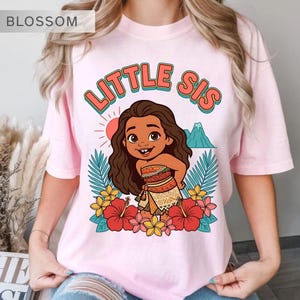 May include: Pink t-shirt with the words "LITTLE SIS" in a retro font, featuring a cartoon illustration of a Polynesian girl surrounded by flowers, palm leaves, a mountain, and a sun.