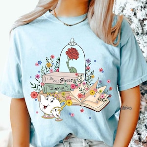 Puede incluir: Una camiseta azul claro con un estampado gráfico de una rosa roja en una cúpula de cristal, una pila de libros con el texto Be our Guest y Tale as old as Time, una tetera con una taza y un libro abierto rodeado de ilustraciones florales.