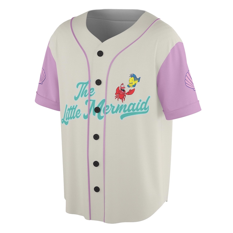May include: Cream-colored baseball jersey with light purple sleeves and trim. Features the text "The Little Mermaid" in teal, along with cartoon images of Sebastian and Flounder. A seashell graphic is on the sleeves.