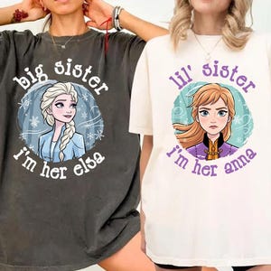 May include: Two t-shirts, one dark gray and one off-white, featuring cartoon images of Elsa and Anna from Frozen. The dark gray shirt says "Big Sister, I'm her Elsa." The off-white shirt says "Lil' Sister, I'm her Anna."