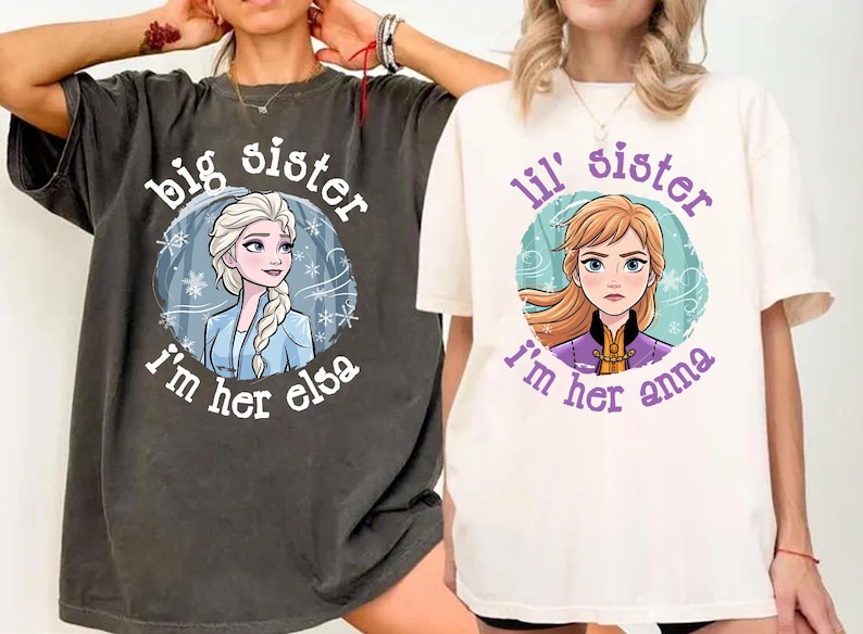 May include: Two t-shirts, one dark gray and one off-white, featuring cartoon images of Elsa and Anna from Frozen. The dark gray shirt says "Big Sister, I'm her Elsa." The off-white shirt says "Lil' Sister, I'm her Anna."