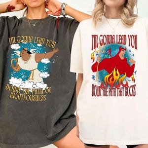 Puede incluir: Dos camisetas gráficas. Una es gris oscuro con una ilustración de un personaje y el texto "I'M GONNA LEAD YOU DOWN THE PATH OF RIGHTEOUSNESS". La otra es blanquecina con un gráfico de diablo y el texto "I'M GONNA LEAD YOU DOWN THE PATH THAT ROCKS".