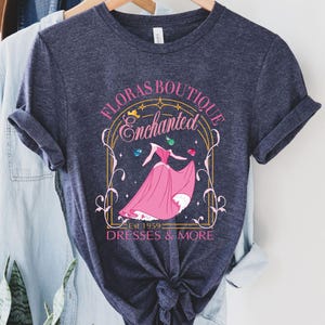 May include: A navy blue t-shirt with a gold frame design featuring a princess in a pink dress and the text "Floras Boutique Enchanted Est 1959 Dresses & More".