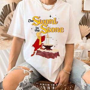 K&ouml;nnte beinhalten: Wei&szlig;es T-Shirt mit einem Motiv aus "The Sword in the Stone". Das Design zeigt eine Figur in roter und gelber Kleidung, die ein Schwert aus einem Amboss zieht. Der Text "The Sword in the Stone" ist gelb und lila.