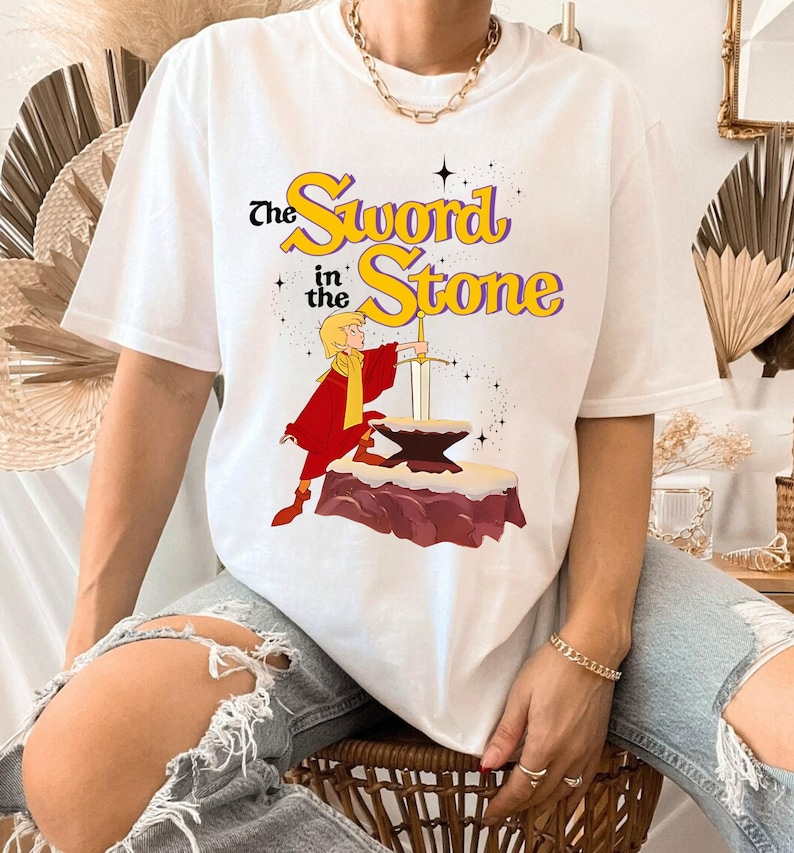K&ouml;nnte beinhalten: Wei&szlig;es T-Shirt mit einem Motiv aus "The Sword in the Stone". Das Design zeigt eine Figur in roter und gelber Kleidung, die ein Schwert aus einem Amboss zieht. Der Text "The Sword in the Stone" ist gelb und lila.