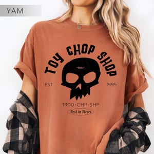 Pu&ograve; includere: Una t-shirt a maniche corte color ruggine con una grafica nera raffigurante un teschio. Il testo recita TOY CHOP SHOP, EST. 1995, 1800-CHP-SHP e Rest in Pieces. La t-shirt &egrave; indossata con una camicia di flanella a quadri bianchi e neri.
