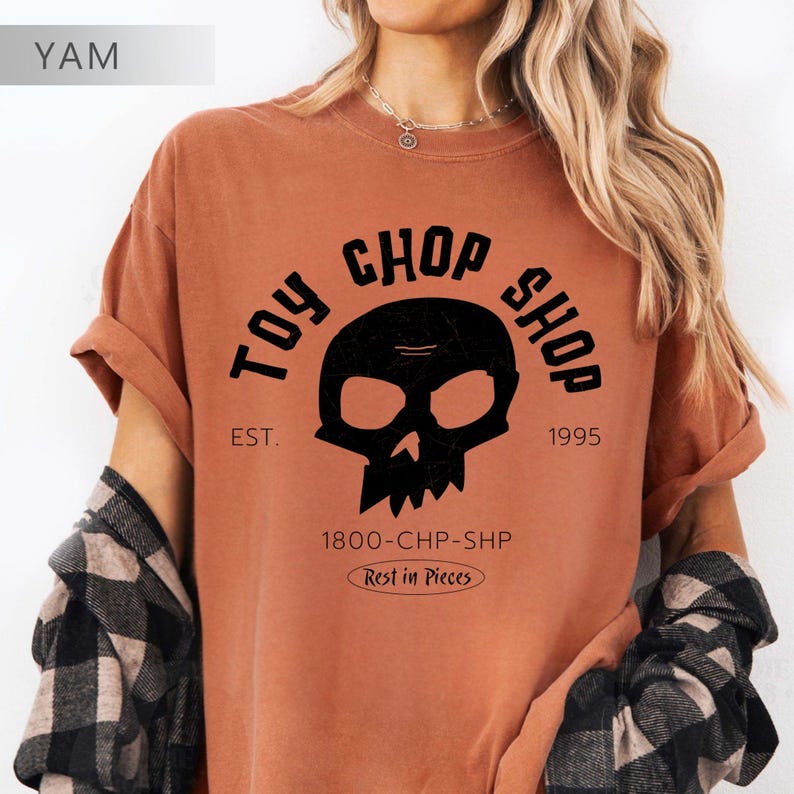 Pu&ograve; includere: Una t-shirt a maniche corte color ruggine con una grafica nera raffigurante un teschio. Il testo recita TOY CHOP SHOP, EST. 1995, 1800-CHP-SHP e Rest in Pieces. La t-shirt &egrave; indossata con una camicia di flanella a quadri bianchi e neri.