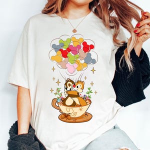 May include: White t-shirt with a cartoon image of Chip and Dale from Disney's Chip 'n Dale Rescue Rangers. The characters are holding onto a teacup with a floral design and are surrounded by colorful Mickey Mouse balloons.