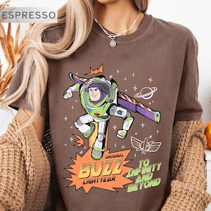 K&ouml;nnte beinhalten: Espressofarbenes T-Shirt mit einer Grafik von Buzz Lightyear in seinem Raumanzug. Das Design zeigt den Text "Original Buzz Lightyear" und "To Infinity and Beyond" in Orange und Gr&uuml;n. Das T-Shirt hat einen Rundhalsausschnitt.