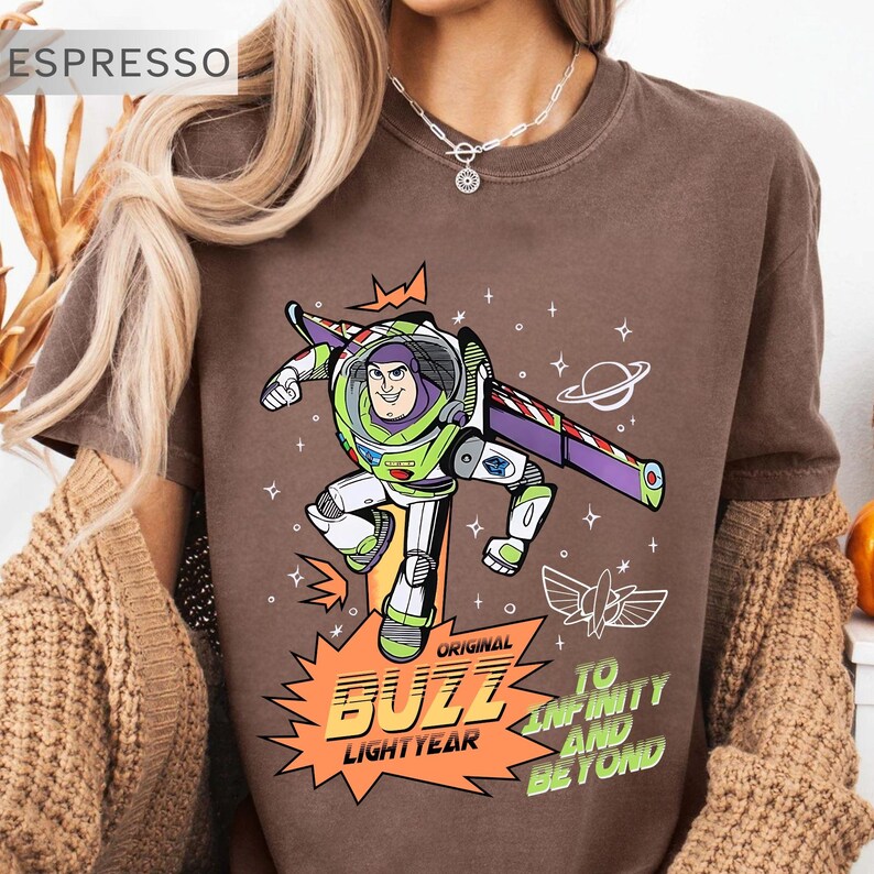 K&ouml;nnte beinhalten: Espressofarbenes T-Shirt mit einer Grafik von Buzz Lightyear in seinem Raumanzug. Das Design zeigt den Text "Original Buzz Lightyear" und "To Infinity and Beyond" in Orange und Gr&uuml;n. Das T-Shirt hat einen Rundhalsausschnitt.