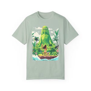 Puede incluir: Una camiseta verde claro con un estampado gr&aacute;fico de una figura de diosa verde con una mujer de pie frente a ella. La diosa est&aacute; rodeada de exuberante vegetaci&oacute;n verde y flores. La mujer sostiene una lanza y lleva una falda marr&oacute;n y una blusa roja.