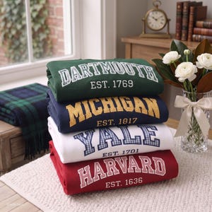 May include: A stack of four folded sweatshirts in green, navy blue, white, and red. Each sweatshirt features a university name in a collegiate font with a plaid pattern fill and an established year printed underneath: Dartmouth 1769, Michigan 1817, Yale 1701, and Harvard 1636.