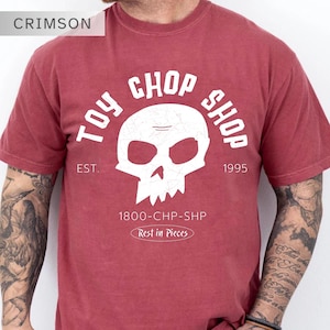 Pu&ograve; includere: Una t-shirt a maniche corte color cremisi con una grafica bianca raffigurante un teschio. Il testo recita TOY CHOP SHOP, EST. 1995, 1800-CHP-SHP e Rest in Pieces. Il design ha un aspetto usurato e invecchiato con una texture screpolata.