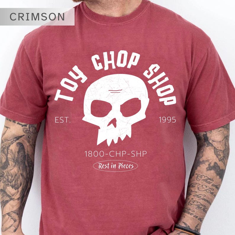 Pu&ograve; includere: Una t-shirt a maniche corte color cremisi con una grafica bianca raffigurante un teschio. Il testo recita TOY CHOP SHOP, EST. 1995, 1800-CHP-SHP e Rest in Pieces. Il design ha un aspetto usurato e invecchiato con una texture screpolata.