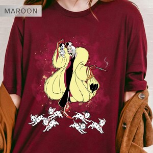 K&ouml;nnte beinhalten: Burgunderfarbenes Langarmshirt mit einer Cartoon-Illustration einer Figur in einem gelben Pelzmantel, einem schwarzen Kleid und einem kleinen Hund. Das Shirt zeigt auch mehrere Dalmatiner-Welpen. Das Wort "MAROON" befindet sich oben links.