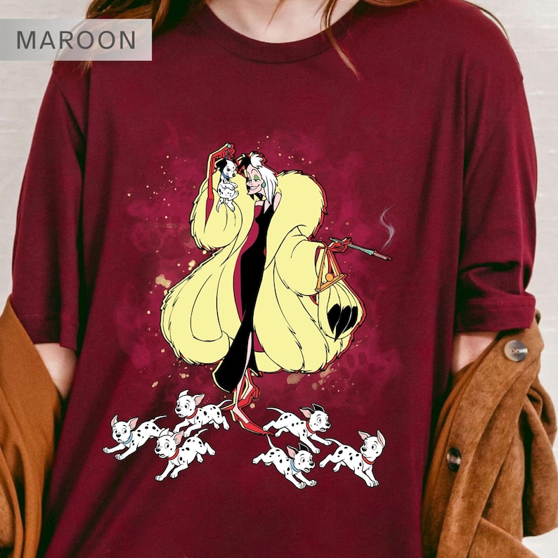 K&ouml;nnte beinhalten: Burgunderfarbenes Langarmshirt mit einer Cartoon-Illustration einer Figur in einem gelben Pelzmantel, einem schwarzen Kleid und einem kleinen Hund. Das Shirt zeigt auch mehrere Dalmatiner-Welpen. Das Wort "MAROON" befindet sich oben links.
