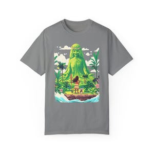 Puede incluir: Una camiseta gris con un estampado gr&aacute;fico colorido de una diosa verde con una corona de flores, de pie con los brazos extendidos. Una peque&ntilde;a figura con una lanza se encuentra frente a la diosa, mir&aacute;ndola.