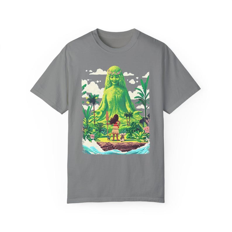 Puede incluir: Una camiseta gris con un estampado gr&aacute;fico colorido de una diosa verde con una corona de flores, de pie con los brazos extendidos. Una peque&ntilde;a figura con una lanza se encuentra frente a la diosa, mir&aacute;ndola.