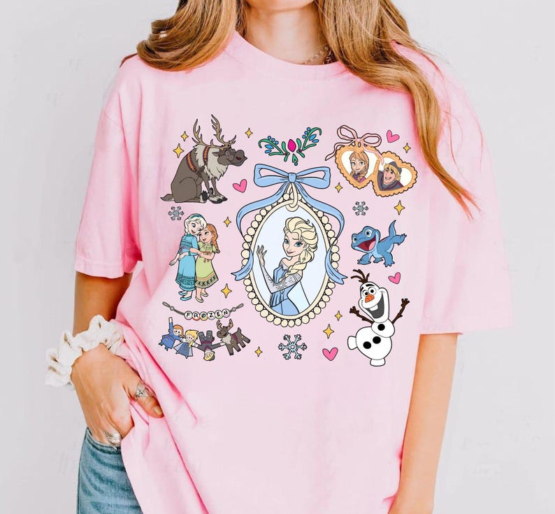 Puede incluir: Camiseta rosa claro con un dise&ntilde;o gr&aacute;fico colorido con personajes de dibujos animados. El dise&ntilde;o incluye ilustraciones de un reno, un mu&ntilde;eco de nieve y dos princesas, junto con otros elementos.