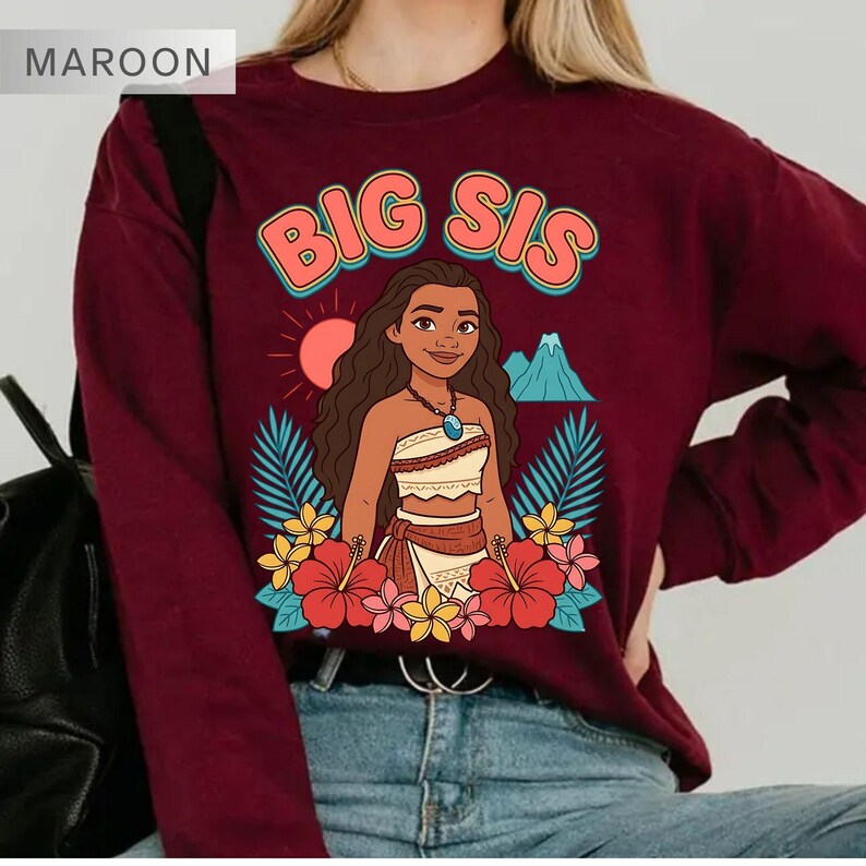 May include: Maroon sweatshirt with a graphic of a cartoon character and the text "BIG SIS". The graphic includes a sun, mountain, and flowers. The sweatshirt is a solid maroon color.