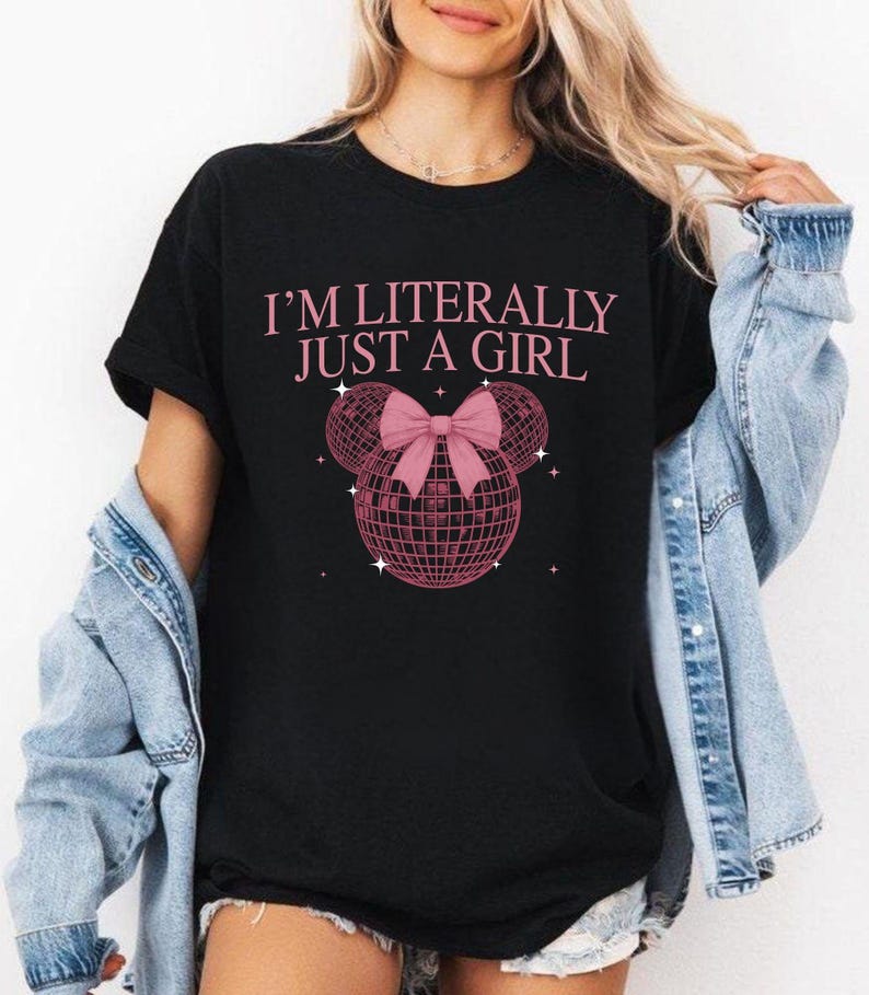 May include: Black t-shirt with the text "I'M LITERALLY JUST A GIRL" in pink. The design includes a pink bow and two disco ball-shaped spheres. The shirt is short-sleeved and appears to be made of a soft material.