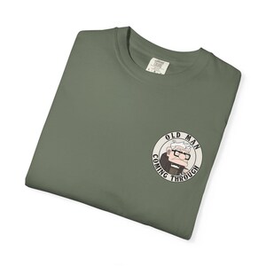 May include: Olive green t-shirt with a circular graphic of an elderly man wearing glasses and the text "OLD MAN COMING THROUGH". The shirt is folded and has a tag in the upper back.