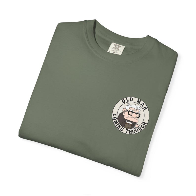 May include: Olive green t-shirt with a circular graphic of an elderly man wearing glasses and the text "OLD MAN COMING THROUGH". The shirt is folded and has a tag in the upper back.