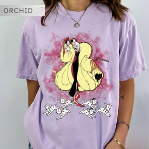 K&ouml;nnte beinhalten: Helllila T-Shirt mit einer Cartoon-Illustration einer Figur in einem gelben Mantel und schwarzen Kleid, die einen kleinen Hund und eine Handtasche h&auml;lt, mit mehreren Dalmatiner-Welpen, die darunter laufen. Das Wort "ORCHID" befindet sich in der oberen linken Ecke.