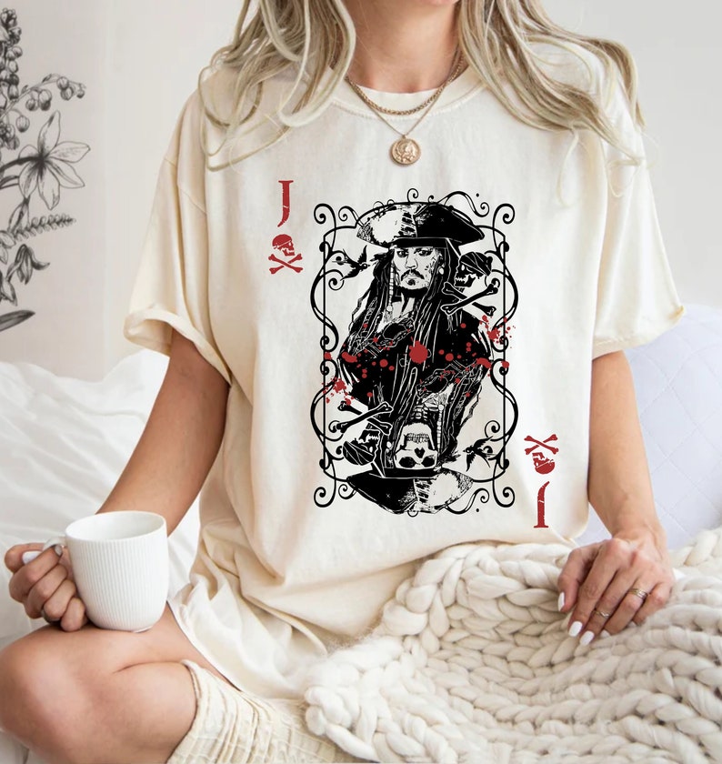 May include: Cream-colored t-shirt featuring a black and red graphic of a playing card with a pirate design. The card shows a pirate with a hat, skulls, and crossbones. The text "J" and "I" are visible.