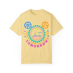 May include: A pale yellow short-sleeve t-shirt featuring a graphic of a futuristic building inside a gear-shaped frame. The text "THERE'S A GREAT, BIG, BEAUTIFUL TOMORROW" is printed in pink and orange, surrounded by blue and orange gear icons.