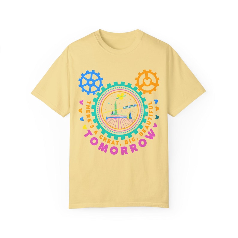 May include: A pale yellow short-sleeve t-shirt featuring a graphic of a futuristic building inside a gear-shaped frame. The text "THERE'S A GREAT, BIG, BEAUTIFUL TOMORROW" is printed in pink and orange, surrounded by blue and orange gear icons.